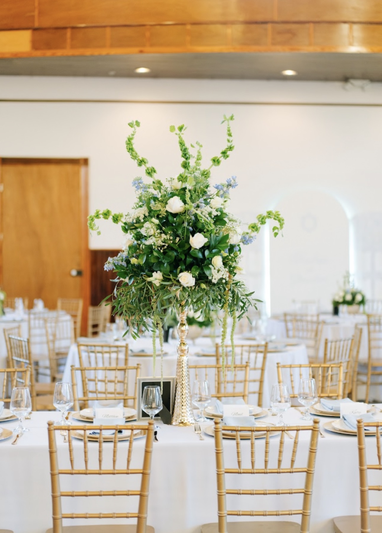 Indoor reception table setup with white chiavari chairs, tall floral centerpiece, and green drapery accents.